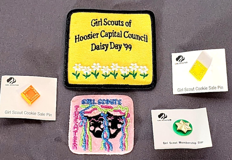May include: A collection of Girl Scout memorabilia, including patches and pins. A yellow patch with black trim reads "Girl Scouts of Hoosier Capital Council Daisy Day '99." Other items include a pink patch and cookie sale pins.