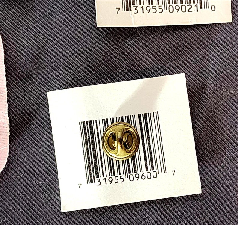 May include: A small, gold-colored lapel pin with a circular design is displayed on a white tag with a barcode and the number "31955 09600 7". The pin is set against a dark gray textured background.