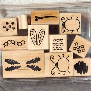 May include: A set of ten wooden rubber stamps housed in a clear plastic storage tray. Each light-colored wooden block features a unique black ink design, including various insects, stars, swirls, wavy lines, and abstract shapes, with 2002 Stampin' Up! text.