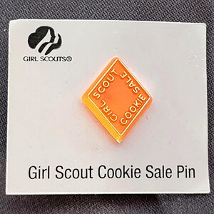 May include: A Girl Scout Cookie Sale pin. The pin is orange and diamond-shaped, with the words "Girl Scout Cookie Sale" around the edges. The pin is on a white card with the Girl Scouts logo and the text "Girl Scout Cookie Sale Pin".
