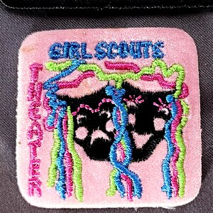 May include: A pink Girl Scouts patch with embroidered text and a black graphic. The patch features the words "GIRL SCOUTS" and "MACARTHUR" in blue and pink thread, with colorful streamers.