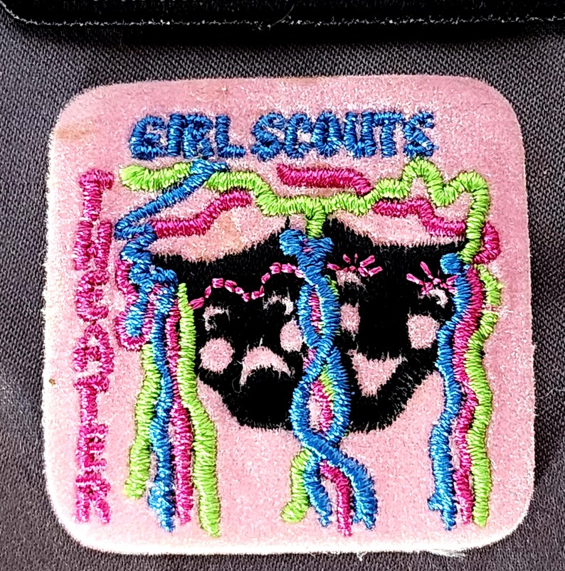 May include: A pink Girl Scouts patch with embroidered text and a black graphic. The patch features the words "GIRL SCOUTS" and "MACARTHUR" in blue and pink thread, with colorful streamers.