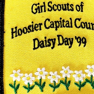May include: A yellow patch with black text that reads "Girl Scouts of Hoosier Capital Cour Daisy Day '99". The bottom of the patch has a row of white and yellow daisy flowers with green stems and leaves.