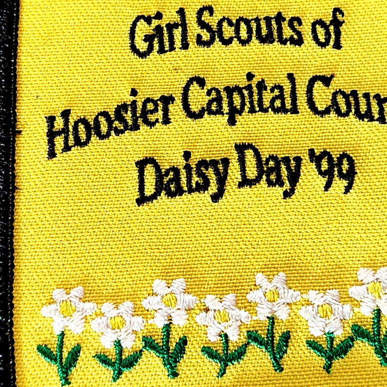 May include: A yellow patch with black text that reads "Girl Scouts of Hoosier Capital Cour Daisy Day '99". The bottom of the patch has a row of white and yellow daisy flowers with green stems and leaves.