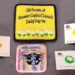 May include: A collection of Girl Scout memorabilia, including patches and pins. A yellow patch reads "Girl Scouts of Hoosier Capital Council Daisy Day '99." Other items include a pink patch and pins labeled "Girl Scout Cookie Sale Pin" and "Girl Scout Membership Star."