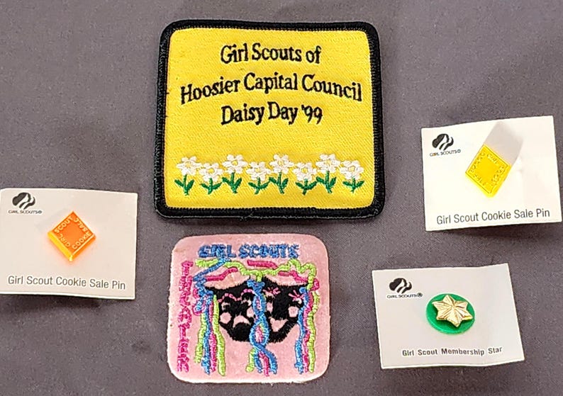 May include: A collection of Girl Scout memorabilia, including patches and pins. A yellow patch reads "Girl Scouts of Hoosier Capital Council Daisy Day '99." Other items include a pink patch and pins labeled "Girl Scout Cookie Sale Pin" and "Girl Scout Membership Star."