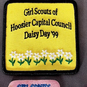 May include: A yellow Girl Scouts patch with black text that reads "Girl Scouts of Hoosier Capital Council Daisy Day '99." The patch has a black border and a row of white daisy flowers with green stems.