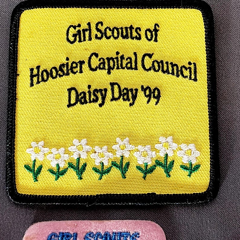 May include: A yellow Girl Scouts patch with black text that reads "Girl Scouts of Hoosier Capital Council Daisy Day '99." The patch has a black border and a row of white daisy flowers with green stems.