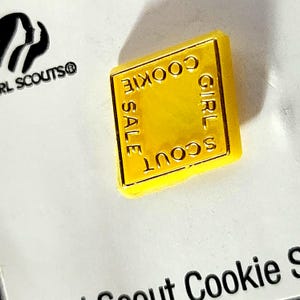 May include: A bright yellow, diamond-shaped pin with the words "GIRL SCOUT COOKIE SALE" in gold lettering. The pin is set against a white background, with the Girl Scouts logo visible.