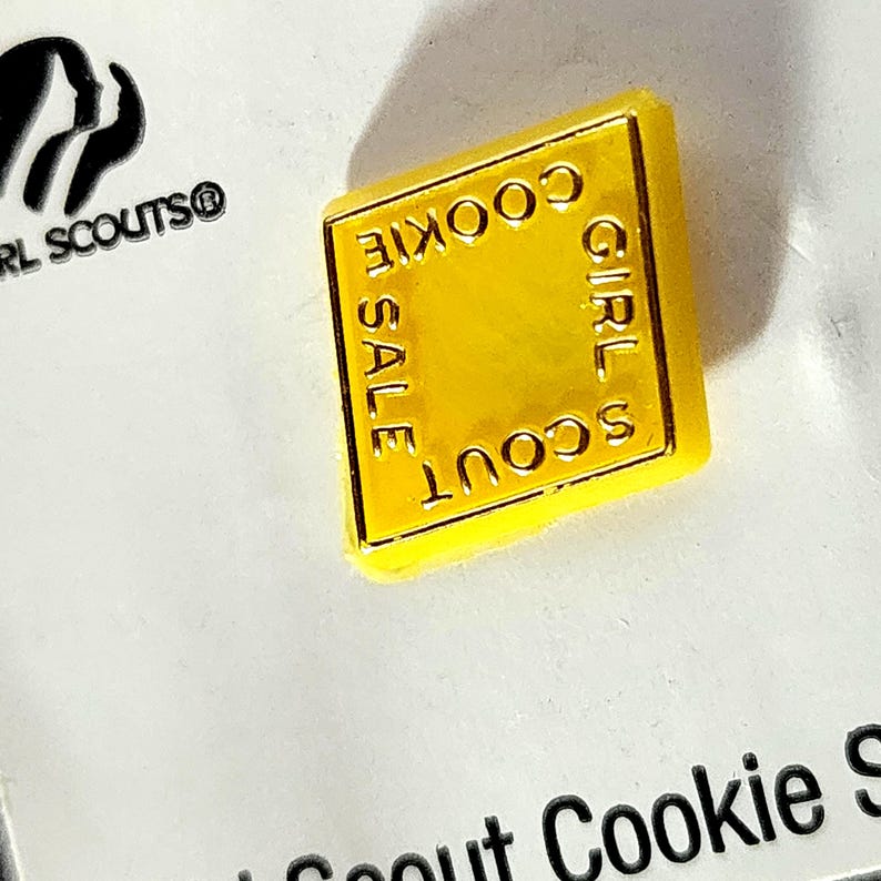 May include: A bright yellow, diamond-shaped pin with the words "GIRL SCOUT COOKIE SALE" in gold lettering. The pin is set against a white background, with the Girl Scouts logo visible.