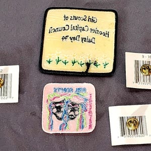 May include: A collection of Girl Scout patches and pins. One patch is yellow with black text and embroidered flowers. Another is pink with a design and text. Three pins are on white cards with barcodes.