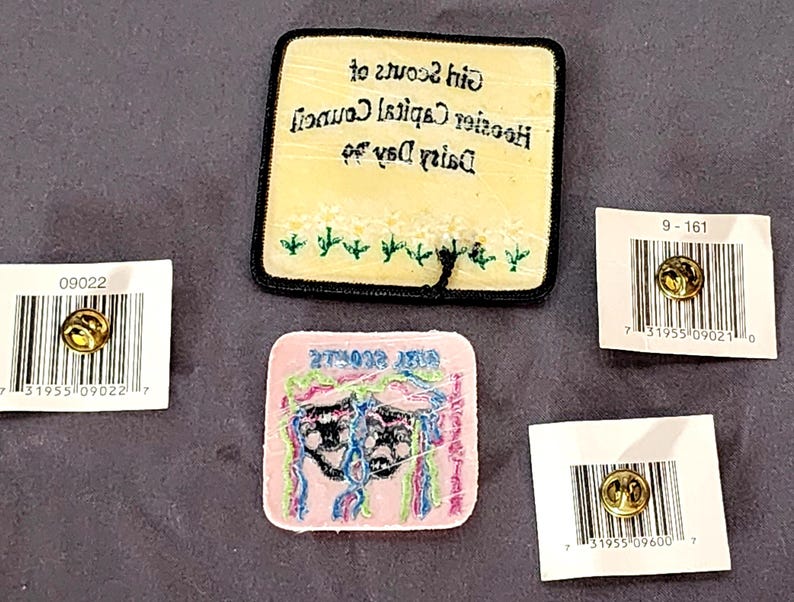 May include: A collection of Girl Scout patches and pins. One patch is yellow with black text and embroidered flowers. Another is pink with a design and text. Three pins are on white cards with barcodes.