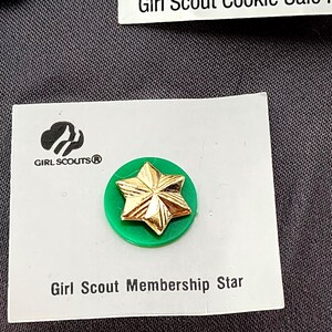 May include: A Girl Scout Membership Star pin. The pin features a gold star on a green circular base, mounted on a white card. The Girl Scouts logo is visible. Text on the card reads "Girl Scout Membership Star".