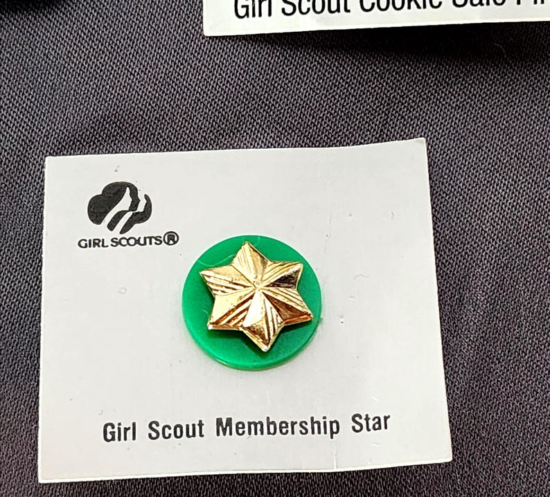 May include: A Girl Scout Membership Star pin. The pin features a gold star on a green circular base, mounted on a white card. The Girl Scouts logo is visible. Text on the card reads "Girl Scout Membership Star".
