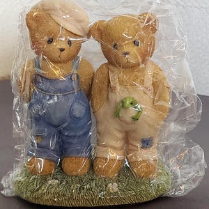 Può includere: Due statuette di orsetti, uno con una salopette blu e un berretto, l'altro con una salopette beige con una rana in tasca. Si trovano su una base erbosa con piccoli fiori bianchi, il tutto racchiuso in plastica.