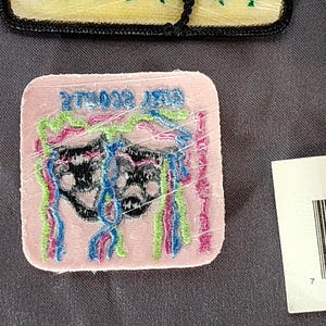 May include: A pink, square patch with the text "BEST SCOUTS EVER" embroidered in green. The patch features a black design with colorful, vertical, fringed accents in blue, pink, and green. The patch is wrapped in clear plastic.