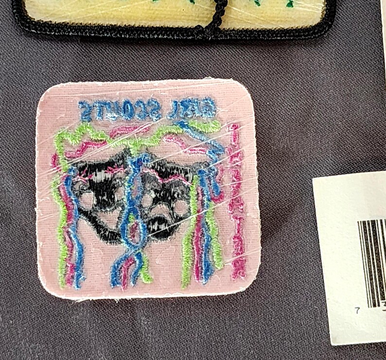 May include: A pink, square patch with the text "BEST SCOUTS EVER" embroidered in green. The patch features a black design with colorful, vertical, fringed accents in blue, pink, and green. The patch is wrapped in clear plastic.