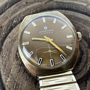 Peut inclure: Montre-bracelet vintage Junghans 17 Jewels avec un boîtier doré et un cadran marron. La montre présente des aiguilles et des index dorés, avec le texte "Junghans 17 Jewels" sur le cadran. Le bracelet est doré et extensible.