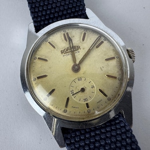 May include: Vintage Roamer Popular watch with a silver case and a cream-colored face. The watch features a small seconds dial and a dark blue fabric band. The watch face displays the word "SWISS".