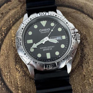 May include: A Casio Quartz wristwatch with a black dial, luminous circular hour markers, and a rotating silver-tone bezel. The watch features a day-date window at the three o'clock position, white hands, and a black rubber strap. Text reads CASIO QUARTZ WATER RESIST 200M.