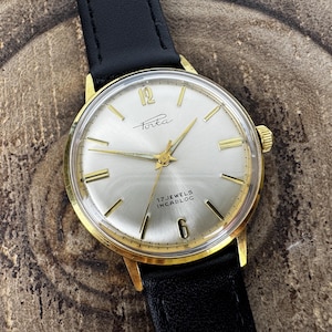 May include: A vintage gold-plated Porta watch with a silver face, gold hands, and hour markers. The watch features a black leather strap. The watch face displays the text "Porta" and "17 Jewels Incabloc". The watch is set against a wooden background.
