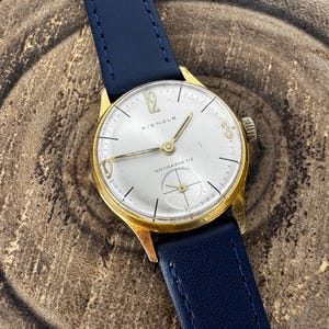 Op de afbeelding: Een vintage goudkleurig Kienzle polshorloge met een zilveren wijzerplaat en een blauwe leren band. De wijzerplaat heeft gouden wijzers en uurmarkeringen, met het woord "ANTIMAGNETIC" op de wijzerplaat. Het horloge ligt op een houten achtergrond.