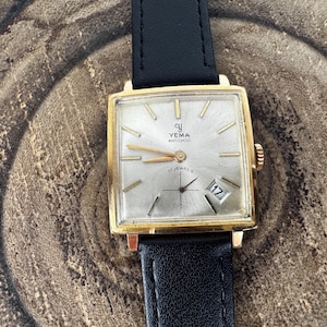 May include: A square gold-tone wristwatch with a silver sunburst dial, gold-tone hands, and gold-tone hour markers. The watch face features the text YEMA, ANTICHOC, and 17 JEWELS, with a date window at the four o'clock position and a black leather strap.