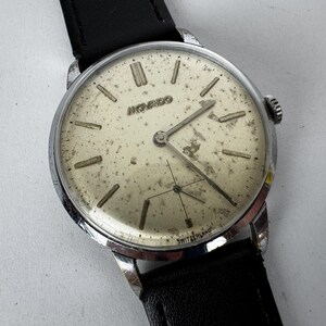 Peut inclure: Montre-bracelet Movado vintage avec un boîtier argenté et un bracelet en cuir noir. Le cadran est blanc cassé avec des index et des aiguilles argentés. Le mot "MOVADO" est imprimé sur le cadran. La montre est marquée "SWITZERLAND".