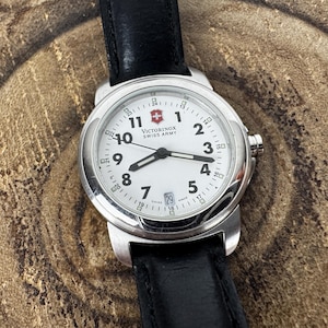 May include: A Victorinox Swiss Army wristwatch with a round silver-tone stainless steel case, a white dial featuring black Arabic numerals, black hour and minute hands, a thin second hand, and a date window at the six o'clock position, attached to a black leather strap.