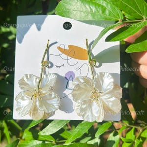 Peut inclure: Une paire de boucles d'oreilles en forme de fleurs nacrées avec des détails dorés. Les boucles d'oreilles sont fixées sur une carte blanche avec le texte "Orangejuicecraft". Le fond est composé de feuilles vertes.