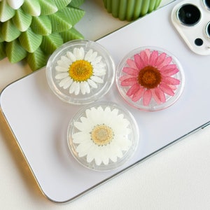 Puede incluir: Tres agarres para teléfono con flores prensadas. Los agarres son transparentes y muestran una margarita blanca, una margarita blanca con un centro amarillo y una margarita rosa con un centro marrón.
