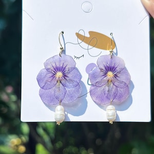 Op de afbeelding: Handgemaakte oorbellen met geperste paarse viooltjes. Elke oorbel heeft een goudkleurige haak en een witte parel. Gepresenteerd op een witte kaart met een minimalistische illustratie.