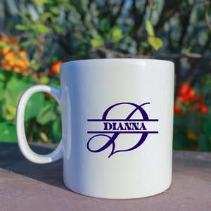 May include: White ceramic coffee mug with a black handle. The mug has a purple  'D' with a line through the middle and the word "Dianna" written on the line.