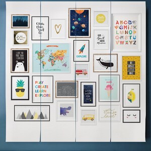 May include: A gallery wall of framed prints with various designs, including a world map, a pineapple wearing sunglasses, a rocket ship, a whale, a school bus, a heart, a lemon, and text prints with phrases like "Oh the Places You'll Go", "Kid You'll Move Mountains", "You Are So Loved", and "Be Brave Little One".