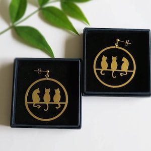 May include: Gold-colored cat silhouette earrings. Each earring features three cat figures perched on a horizontal line within a circular frame. The earrings are displayed in black boxes, with a green leafy branch in the background.