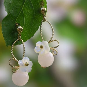 以下が含まれることがあります： 白い花のアクセントとピンクのビーズが付いた、ゴールドのドロップイヤリングのペア。イヤリングは緑の葉からぶら下がっています。
