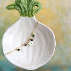 May include: A white ceramic onion-shaped dish with a green leafy top, holding a delicate gold necklace. The necklace features small, colorful beads and a white flower pendant, creating a charming and unique jewelry display.