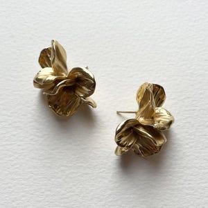 May include: A pair of gold-toned flower-shaped earrings. The earrings are made of metal and have a textured surface.