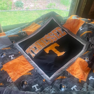 Op de afbeelding: Een patchwork quilt met het logo van de University of Tennessee in de teamkleuren oranje, wit en grijs. Het centrale vierkant toont het woord TENNESSEE in oranje letters boven een grote oranje T, omringd door kleinere vierkanten met teamgraphics.