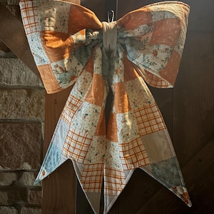 Può includere: Un grande fiocco decorativo realizzato con toppe di tessuto trapuntato che presentano un mix di motivi arancioni, bianchi e azzurri. Il tessuto mostra illustrazioni di zucche, fogliame e disegni a quadri, con due lunghe code appuntite pendenti.
