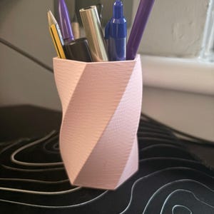 May include: A light pink pen holder with a 3D-printed, twisted design. It contains a yellow pencil, purple pens, a blue marker, and a gold pen. The holder sits on a black surface with a white topographical design.