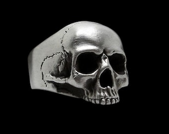 Puede incluir: Un anillo de calavera de plata con detalles realistas. El anillo está hecho de metal y tiene un diseño de calavera realista.