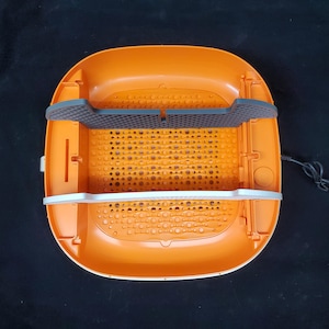 May include: Orange and white plastic food dehydrator with a black mesh tray and a white plastic handle. The dehydrator has a round shape and a textured surface.