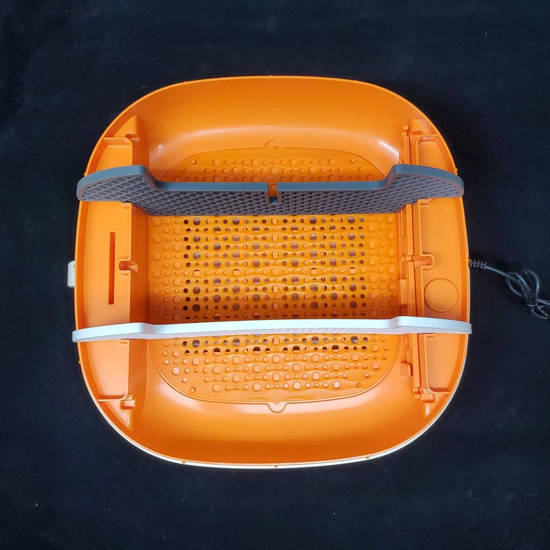 May include: Orange and white plastic food dehydrator with a black mesh tray and a white plastic handle. The dehydrator has a round shape and a textured surface.
