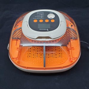 May include: A white and orange automatic egg incubator with a clear plastic lid. The incubator has a digital display and buttons for controlling temperature and humidity. The text "Chickery" is printed on the front of the incubator.