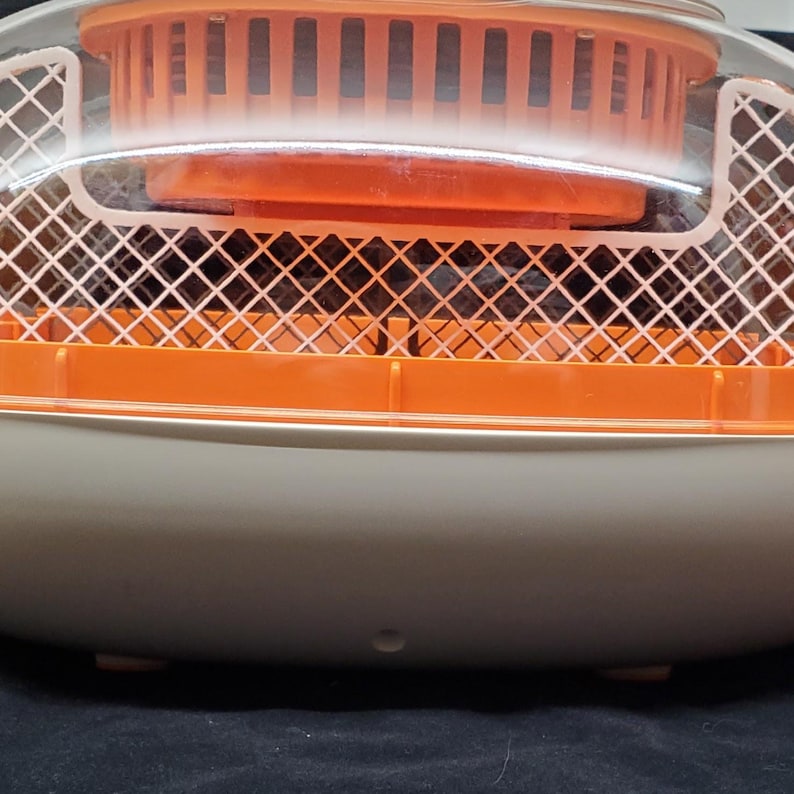 May include: A white and orange plastic egg incubator with a clear lid and a wire mesh basket inside. The incubator has a round base and a small hole in the bottom.