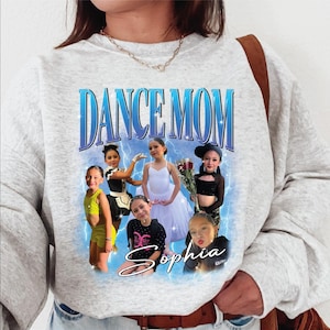 May include: A light gray sweatshirt with a graphic that says "DANCE MOM" in blue text. The graphic features photos of young dancers and the name "Sophia" in cursive. The design includes lightning bolt accents.