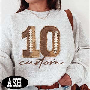 May include: A light gray heathered sweatshirt featuring a large number 10 graphic designed to resemble a football with brown textured panels and white laces. Below the number, the word custom is printed in a brown cursive script font.