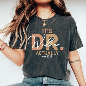 May include: A dark gray t-shirt with the text "It's Dr. Walker Actually est. 2023" in white and peach lettering.