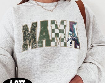 May include: Light gray sweatshirt with the word "MAMA" in a bold, block letter design. The letters have a camouflage, checkered, and marbled pattern. The sweatshirt has a crew neck and long sleeves. The word "ASH" is printed on a black oval at the bottom.